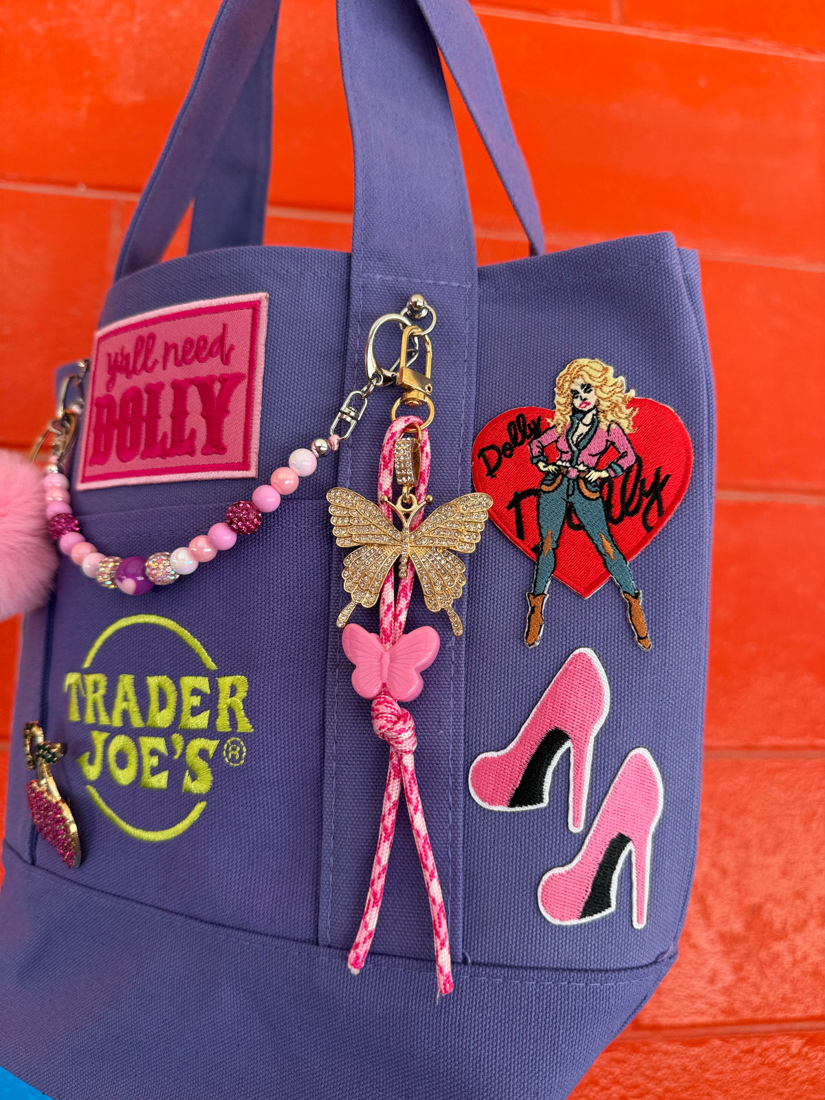 A purple Trader Joe's mini tote with a hot pink 'y'all need DOLLY' patch, the Trader Joe's logo, pink stiletto patches and a butterfly keychain, set against a bright orange brick wall. Dolly Parton Boots & Heels mini tote by PrettyMUCH.