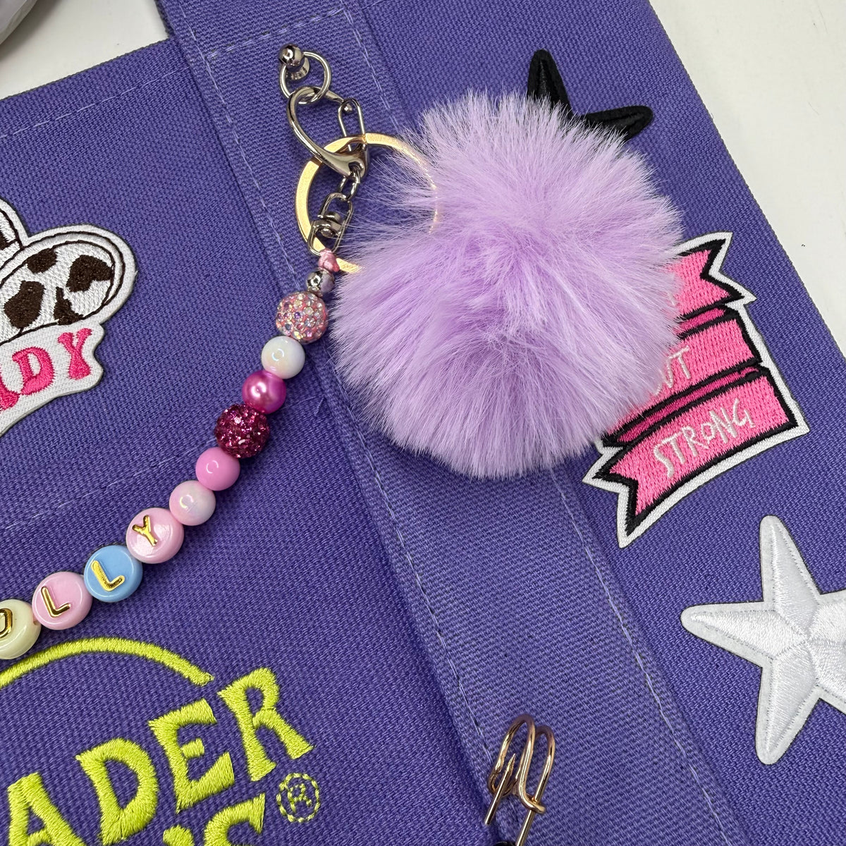 A lavender purple fluffy pom-pom keychain charm with a beaded strap, displayed on a purple canvas tote bag with colorful embroidered patches. Dolly Parton Sweet but Strong mini tote by PrettyMUCH.