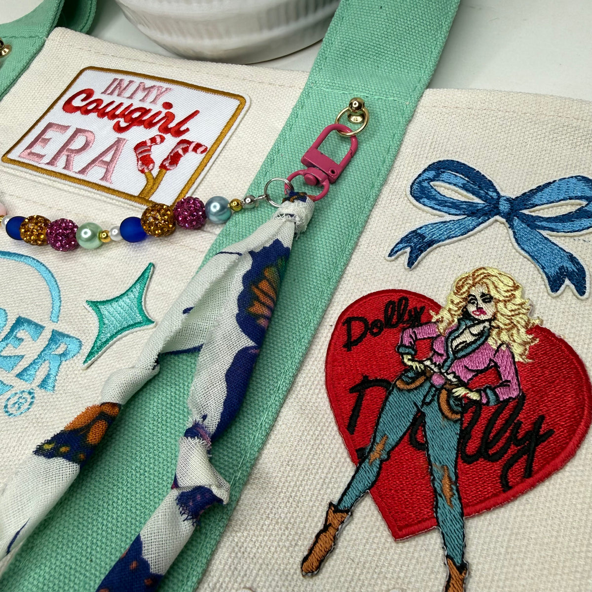 Close-up of a canvas tote bag with embroidered patches and a matching fabric charm. The bag has a mint green strap, a red heart patch with a blonde woman and the words 'Dolly Partly,' a blue bow, and a white patch reading 'In My Cowgirl Era. Dolly Parton Cowgirl Era mini tote by PrettyMUCH.