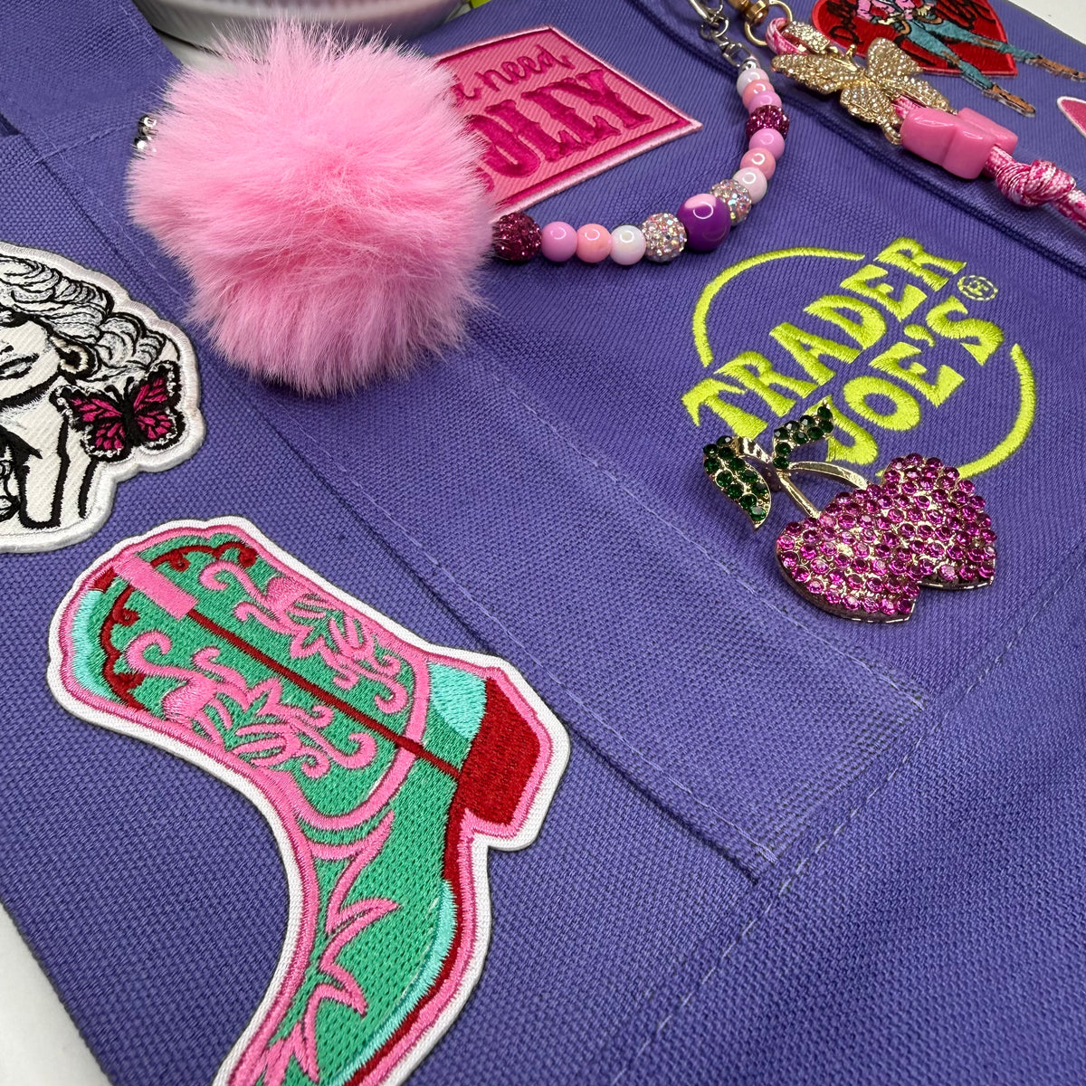 Close-up of the purple Trader Joe's mini tote with an embroidered teal-and-pink cowgirl boot patch, a pink fuzzy pom-pom and a pink-and-purple beaded bag chain. Dolly Parton Boots & Heels mini tote by PrettyMUCH.