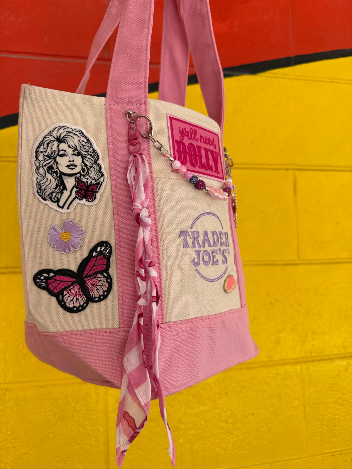 A pink and beige canvas tote bag with embroidered patches, a butterfly applique, and a pink gingham scarf, shown hanging against a bright yellow and red brick wall. Dolly Parton Pink Bandana mini tote by PrettyMUCH.