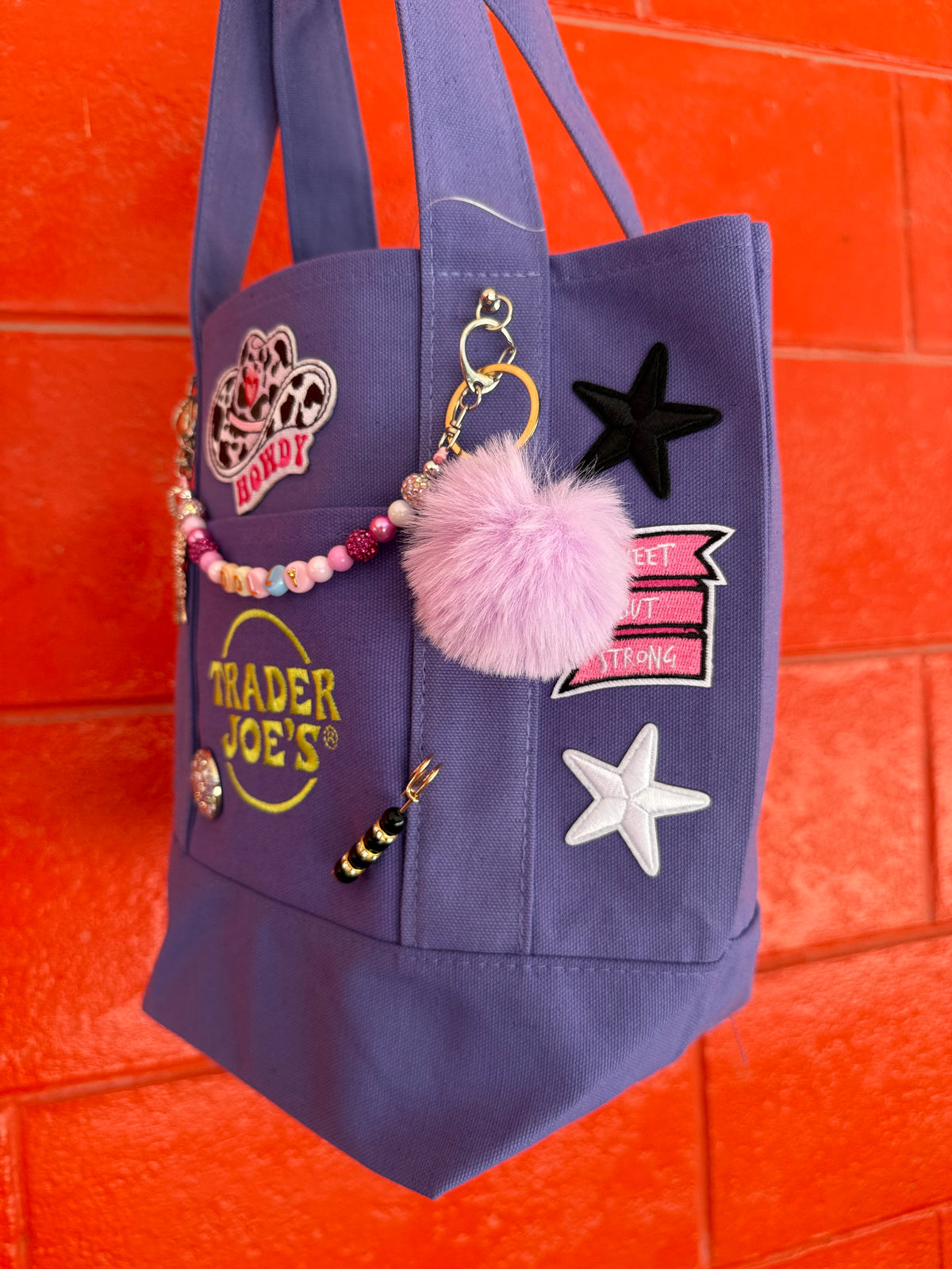 A purple Trader Joe's mini tote decorated with Dolly patches, a row of black and white star patches, a lavender pom-pom and a beaded DOLLY bag chain, hanging against a bright orange brick wall. Dolly Parton Sweet but Strong mini tote by PrettyMUCH.