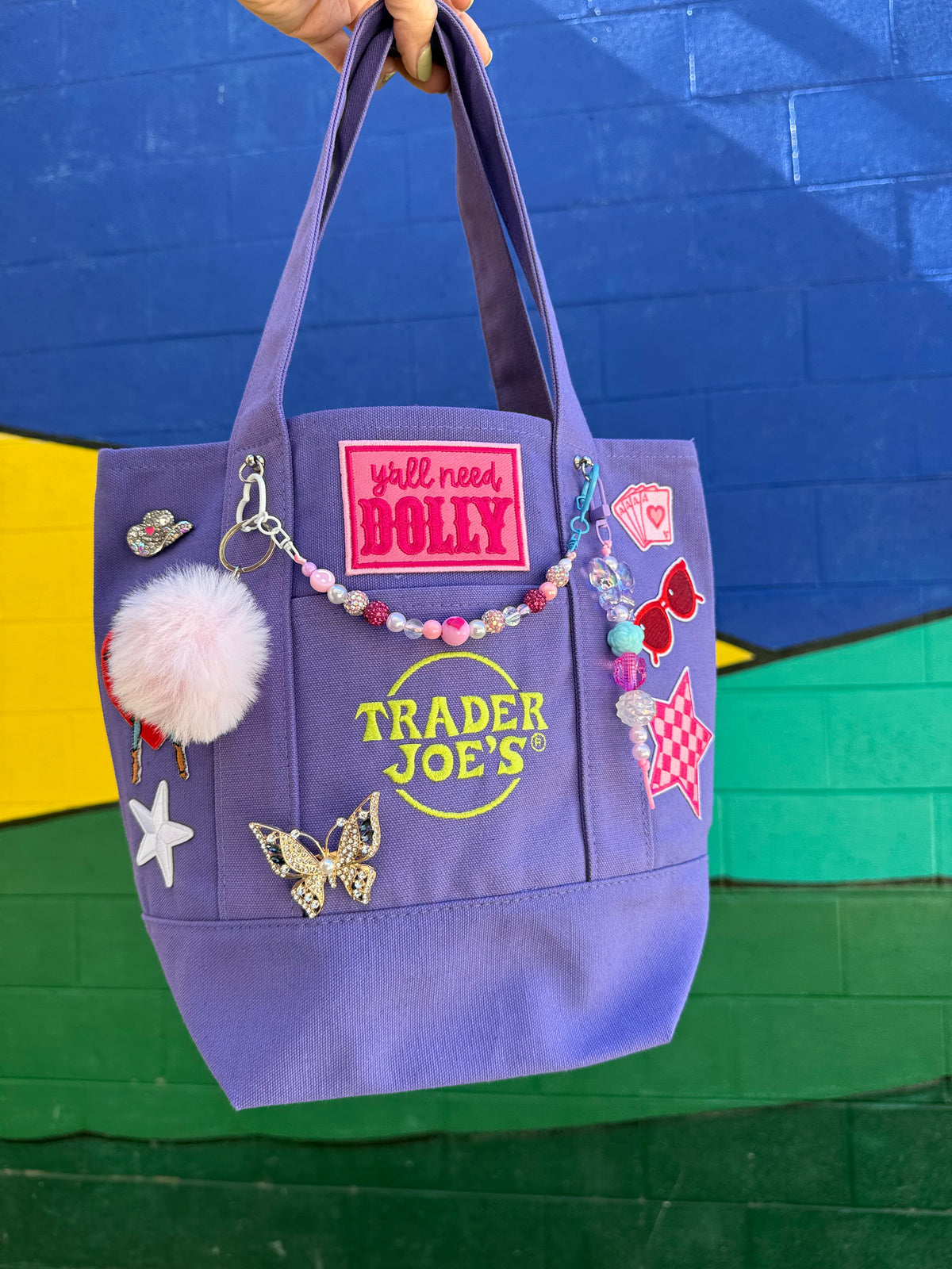 A purple canvas tote bag with 'Trader Joe's' branding, decorated with a pink pom-pom, beaded charm, and various colorful patches, held up by a hand against a painted brick wall. Dolly Parton Aces High mini tote by PrettyMUCH.