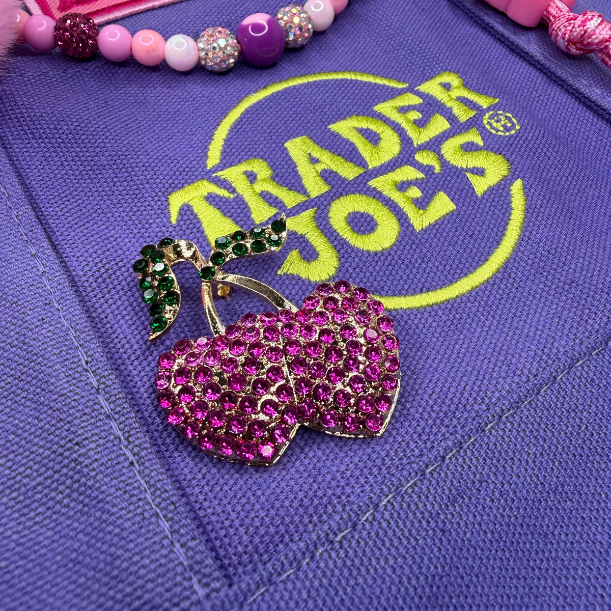 Close-up of the purple Trader Joe's mini tote with the bright yellow Trader Joe's logo and a rhinestone cherry heart brooch pinned on the front, with part of the pink beaded bag chain visible along the top edge. Dolly Parton Boots & Heels mini tote by PrettyMUCH.