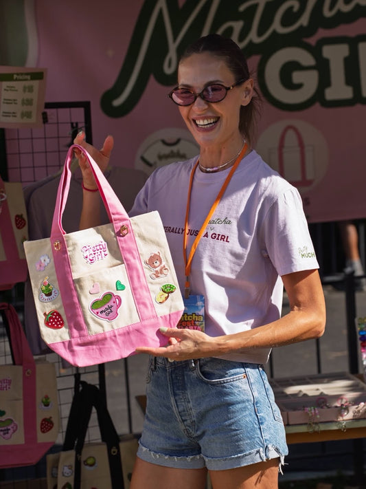 Smiling woman in the Literally Just a Matcha Girl cropped tee and sunglasses holding a pink-trim patch tote at the PrettyMUCH Matcha Fest booth