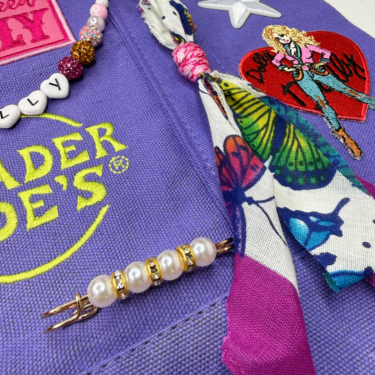 A close-up of a purple denim accessory set featuring a denim hair tie with a colorful butterfly-patterned fabric, a pearl and gold hair clip, and a beaded necklace on a purple denim background. Dolly Parton Y'all Need Dolly mini tote by PrettyMUCH.