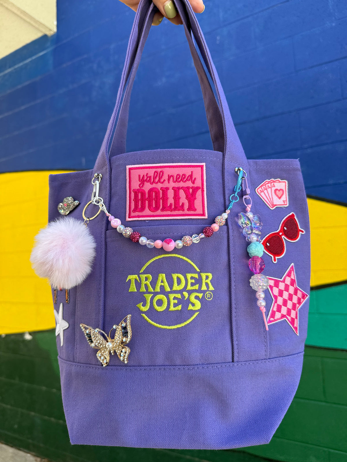 A purple canvas tote bag with 'Trader Joe's' branding, decorated with various colorful patches, a pink pom-pom, and a beaded charm, held up against a colorful painted brick wall. Dolly Parton Aces High mini tote by PrettyMUCH.