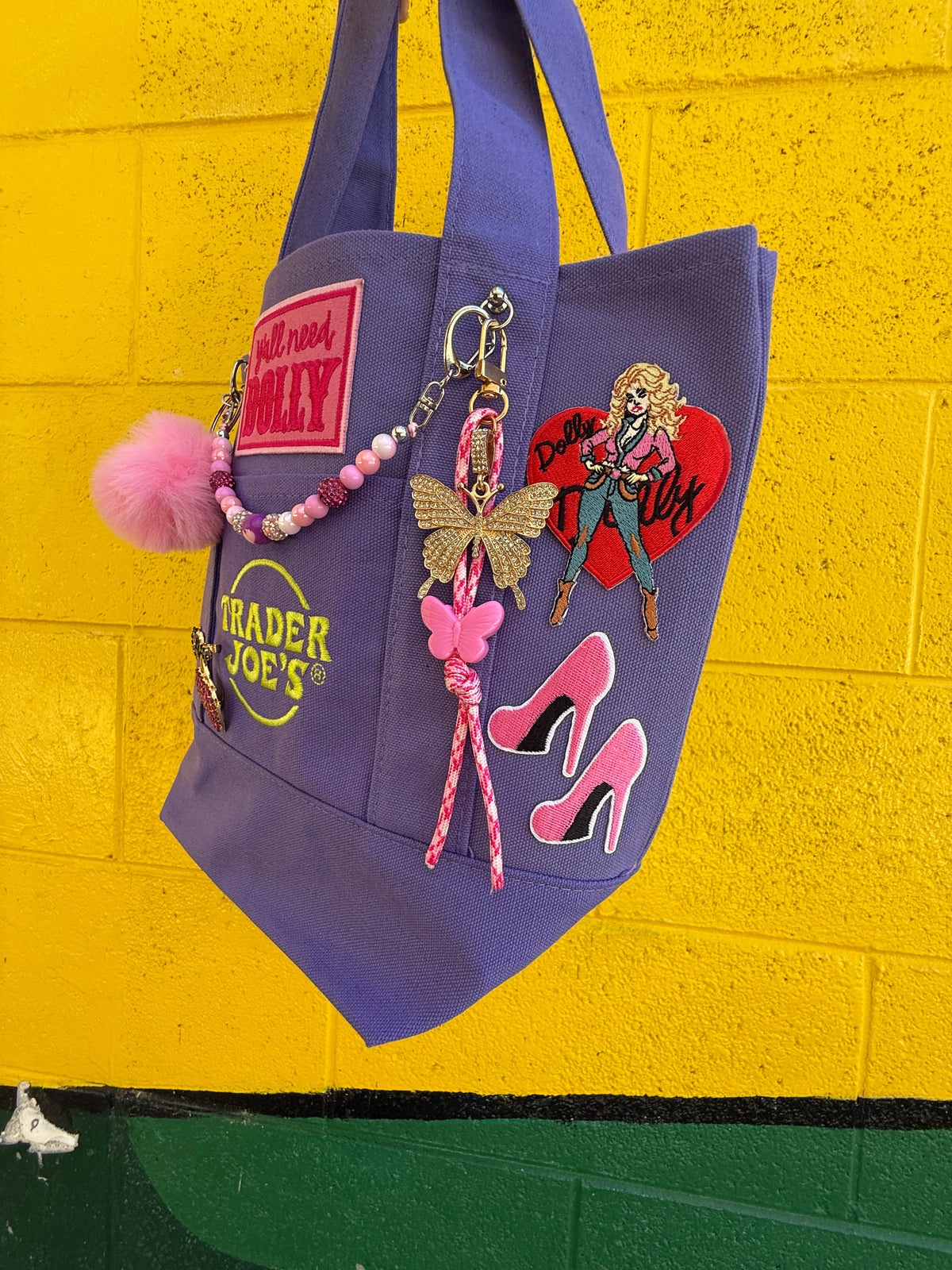 A purple Trader Joe's mini tote with a pink pom-pom, a butterfly charm, pink stiletto patches and the Trader Joe's logo, hanging against a bright yellow brick wall. Dolly Parton Boots & Heels mini tote by PrettyMUCH.
