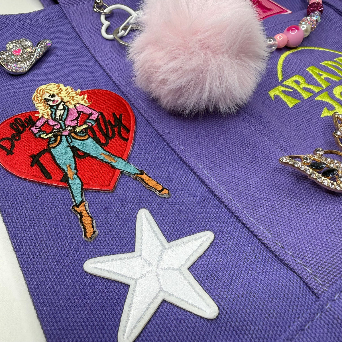 Close-up of the purple Trader Joe's mini tote featuring the embroidered Dolly Parton red heart patch, a white embroidered star, a pink fluffy pom-pom keychain and other accessories. Dolly Parton Aces High mini tote by PrettyMUCH.