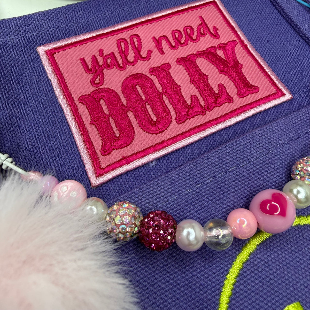 A close-up of a purple fabric item featuring a pink embroidered patch that reads 'y'all need DOLLY' in pink lettering. A beaded strand with pink, pearl, and glittery beads runs across the foreground, with a fluffy white faux-fur trim visible on the left. Dolly Parton Aces High mini tote by PrettyMUCH.