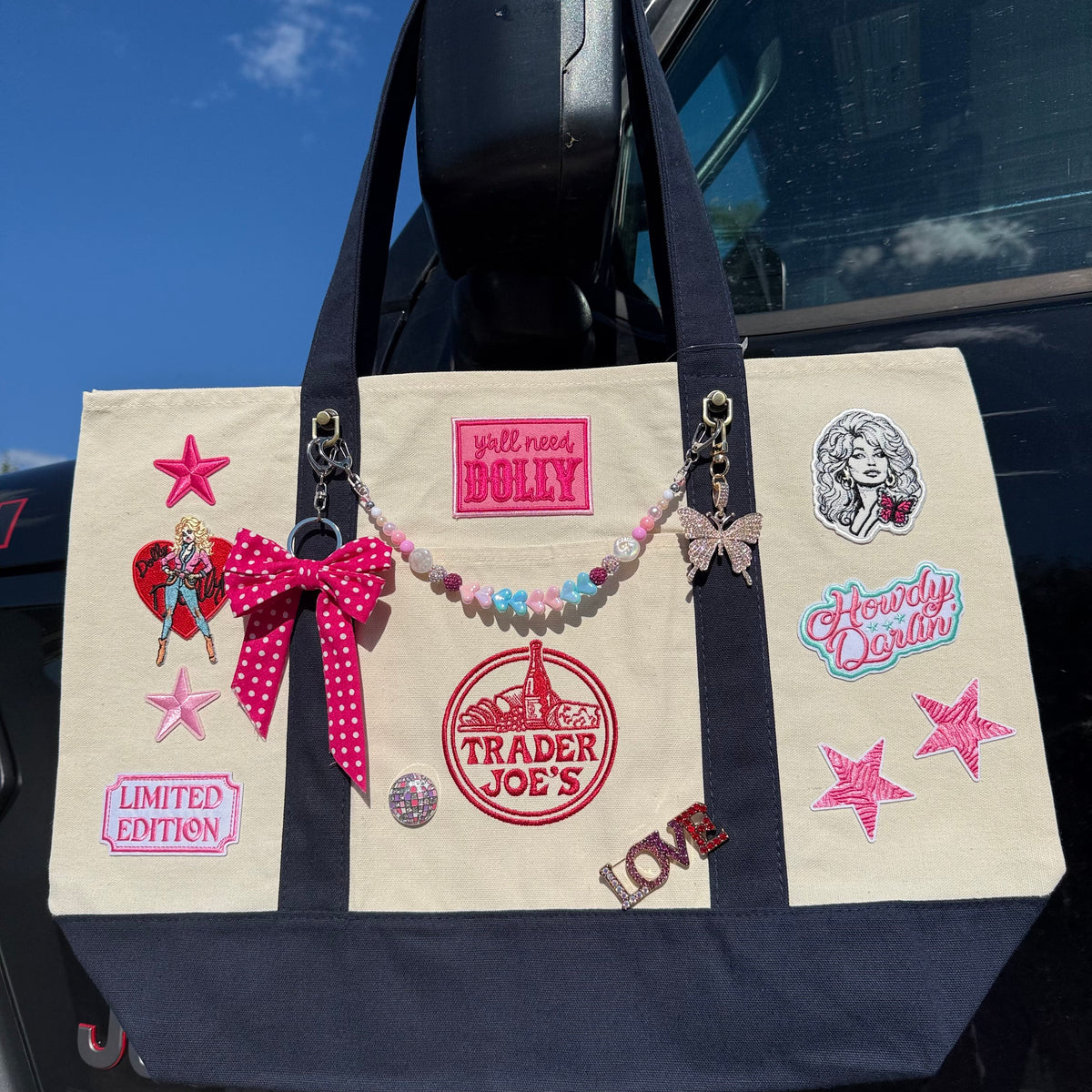 A canvas tote bag with a navy blue bottom and straps, decorated with various pop culture patches, pins, and a beaded charm strap, hanging from a black car door against a blue sky. Dolly Parton Big Love large tote by PrettyMUCH.