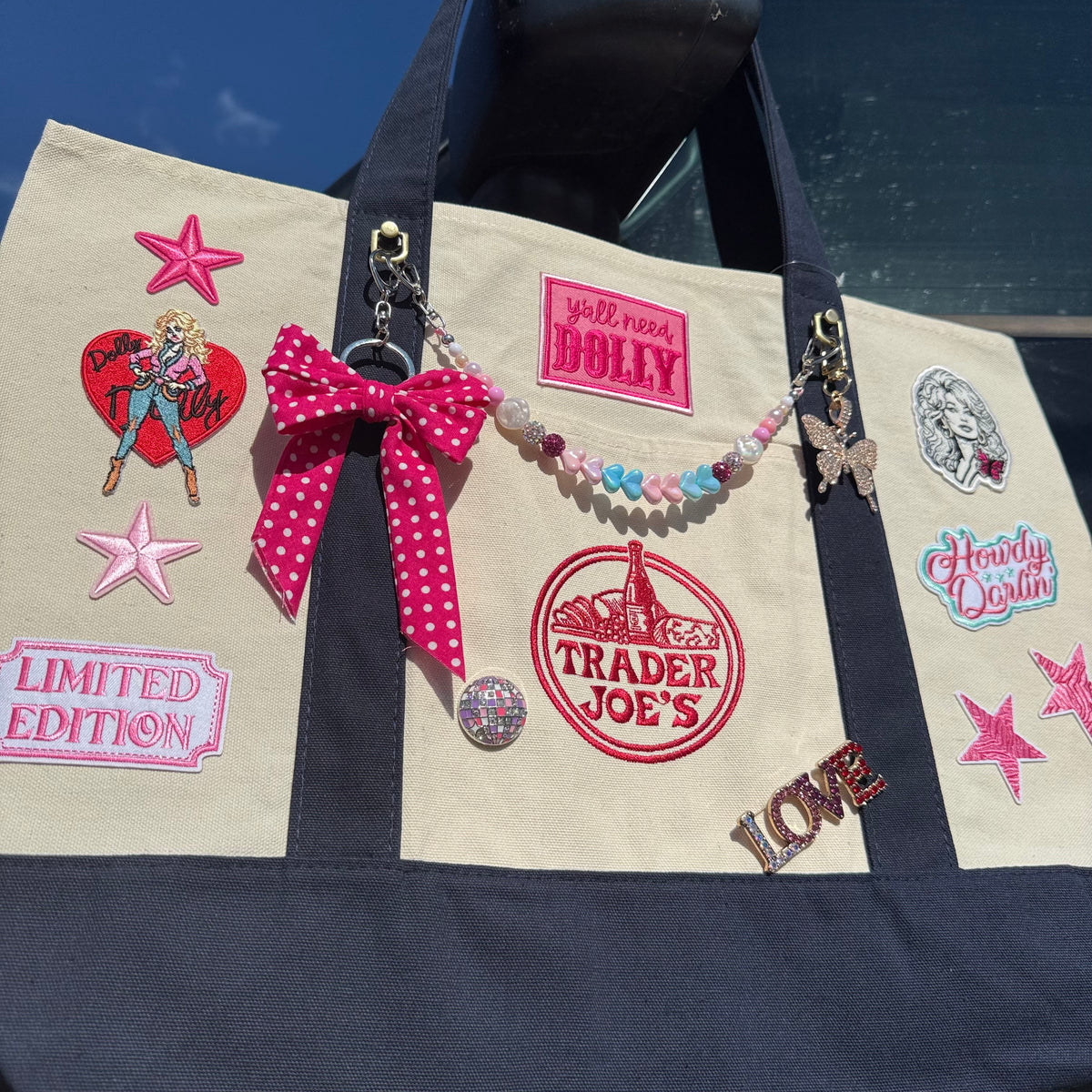 A beige canvas tote bag with navy blue handles and trim, decorated with multiple embroidered patches, a pink polka-dot bow, a beaded charm keychain, and rhinestone pins, photographed outdoors against a blue sky and car window background. Dolly Parton Big Love large tote by PrettyMUCH.