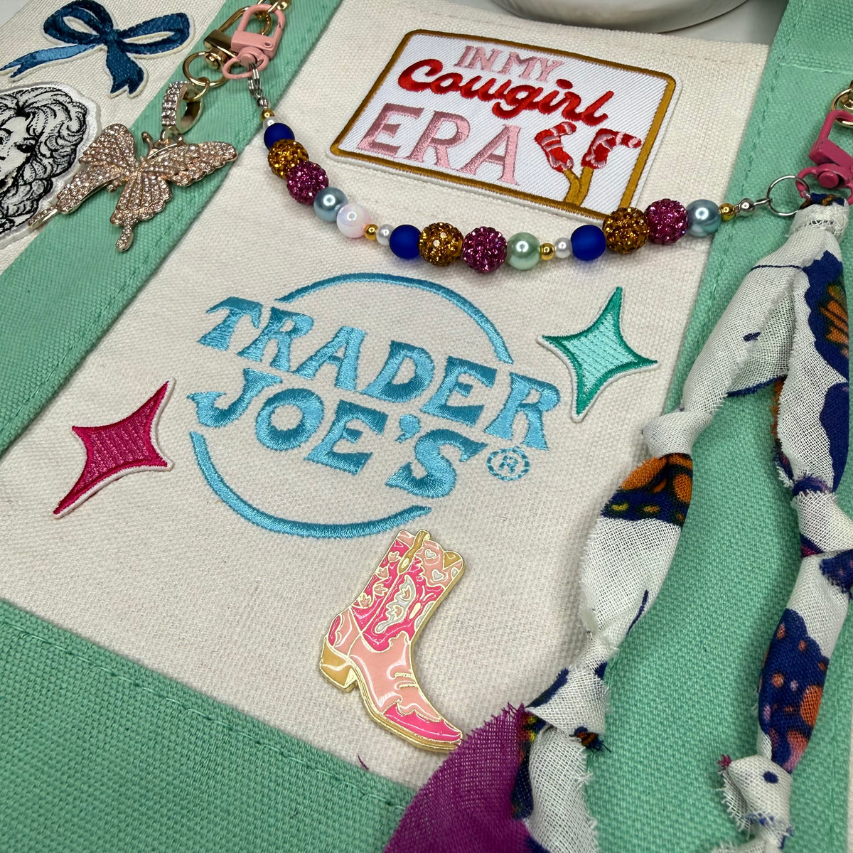 A close-up of a colorful embroidered patch featuring the Trader Joe's logo in blue, surrounded by decorative accessories including a beaded bracelet, a cowboy boot enamel pin, and patterned fabric. Dolly Parton Cowgirl Era mini tote by PrettyMUCH.
