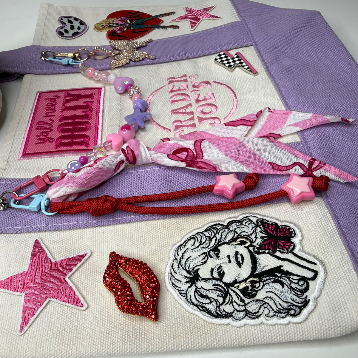 Close-up of the lavender and cream Trader Joe's mini tote with the embroidered Trader Joe's logo, the black-and-white Dolly Parton portrait patch, a pink zebra-stripe star patch, a red rhinestone lips pin and the hot pink 'y'all need DOLLY' patch, with a pink striped bandana twilly and a red cord charm with pink star beads across the front. Dolly Parton Pretty in Pastel mini tote by PrettyMUCH.