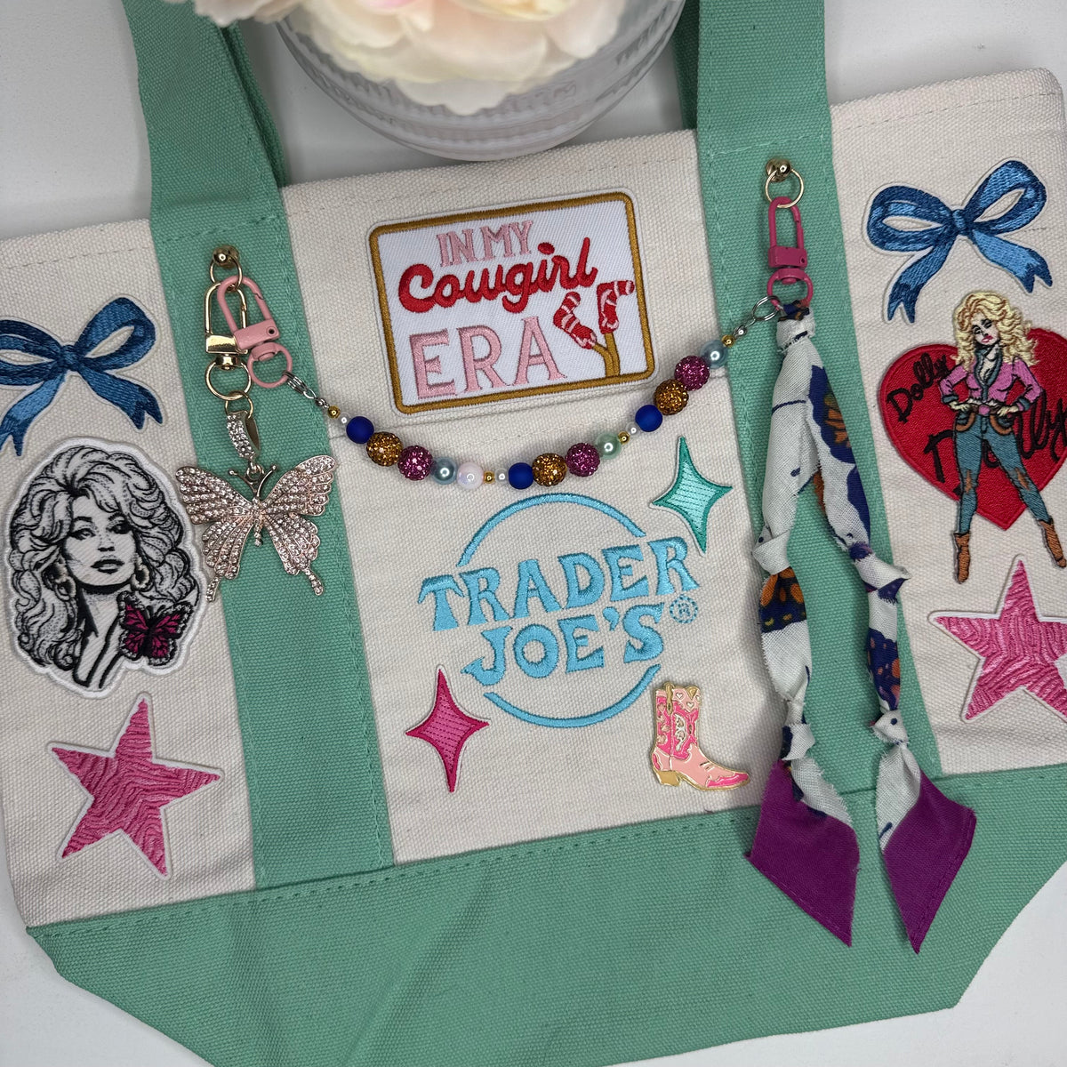 A canvas tote bag with mint green bottom panels and straps, decorated with multiple embroidered patches, appliques, and a beaded keychain. The bag features a central patch reading 'IN MY COWGIRL ERA' and a 'Trader Joe's' logo patch, alongside other western and fashion-themed embellishments. Dolly Parton Cowgirl Era mini tote by PrettyMUCH.