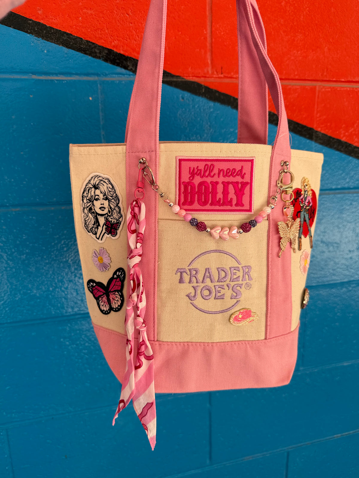 A pink and beige tote bag with multiple decorative patches, charms, and a pink-and-white patterned scarf hanging from the side, set against a bright blue and orange painted brick wall. Dolly Parton Pink Bandana mini tote by PrettyMUCH.