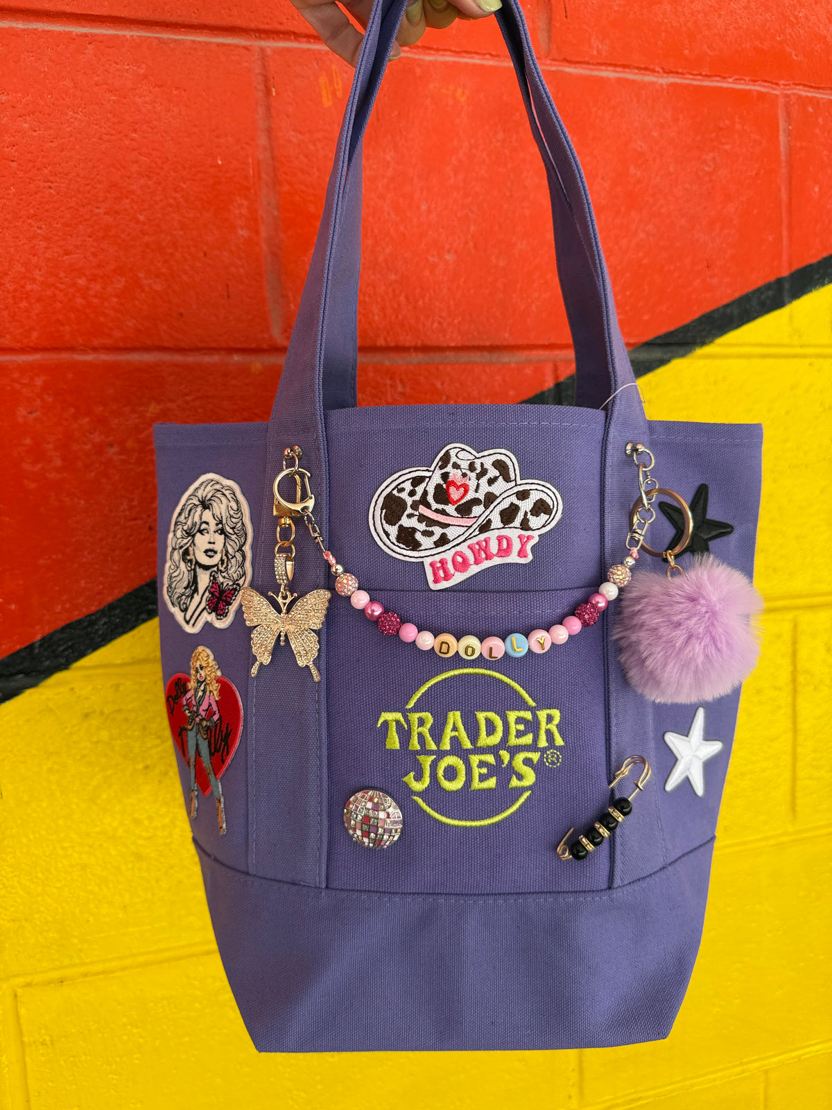 A purple canvas tote bag decorated with various patches, pins, a beaded charm, and a pink pom-pom, featuring the 'Trader Joe's' logo on the front, set against a bright yellow and orange painted brick wall. Dolly Parton Sweet but Strong mini tote by PrettyMUCH.