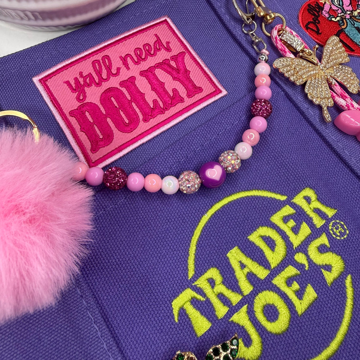 A close-up of a purple fabric item featuring a bright pink embroidered patch that reads 'y'all need DOLLY' and a matching pink beaded charm with a glittery butterfly keychain. A pink pom-pom is also visible on the left, and a Trader Joe's logo is embroidered in yellow-green on the fabric. Dolly Parton Boots & Heels mini tote by PrettyMUCH.
