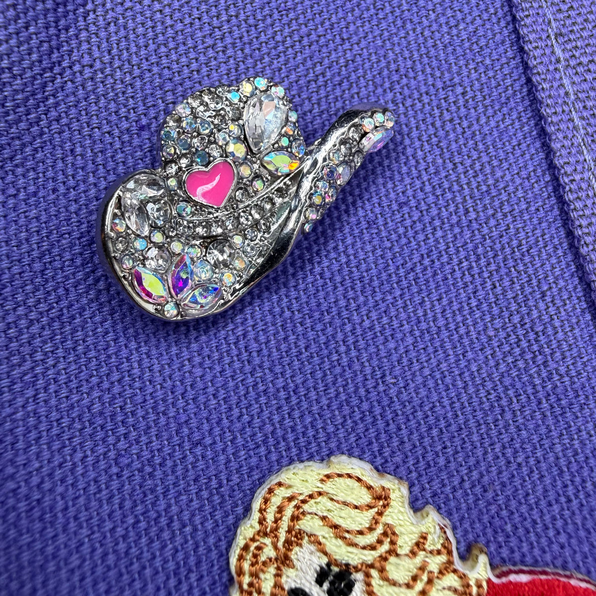 A close-up of a silver-toned rhinestone hand gesture pin with a pink heart, placed on a textured purple garment. Dolly Parton Aces High mini tote by PrettyMUCH.