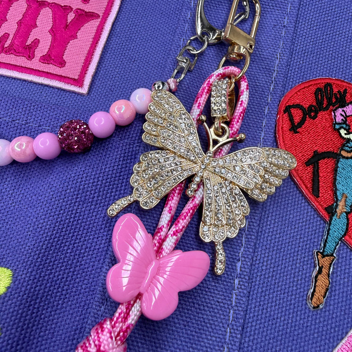 A close-up of a pink and gold keychain or bag charm laid on a blue fabric surface. The charm includes a rhinestone-embellished butterfly, a pink plastic butterfly, a pink braided cord, and pink beads, with a metal clasp at the top. Dolly Parton Boots & Heels mini tote by PrettyMUCH.