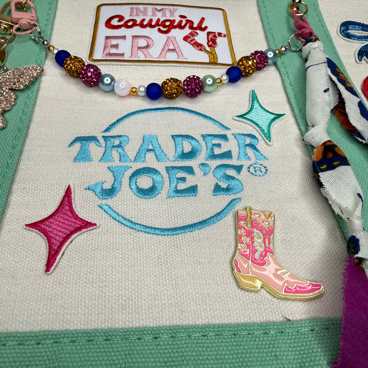 A close-up of a fabric patch featuring the embroidered Trader Joe's logo in light blue, decorated with a pink cowgirl boots pin, a pink glitter star, and a beaded necklace across the top. Dolly Parton Cowgirl Era mini tote by PrettyMUCH.