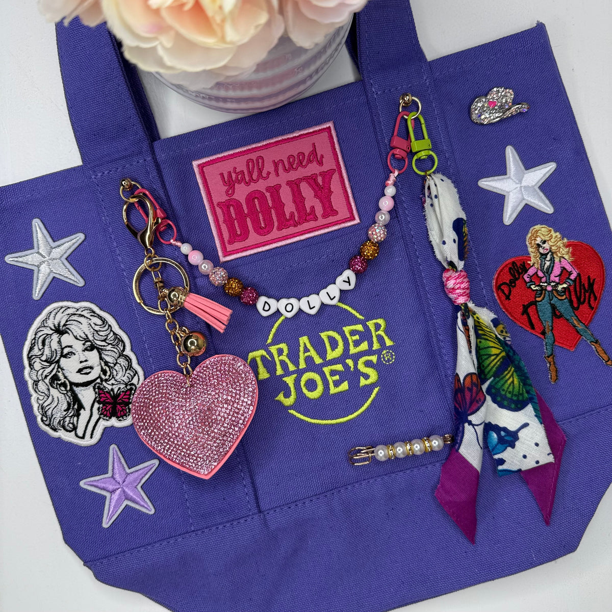 A purple Trader Joe's mini tote decorated with a pink rhinestone heart keychain, a beaded DOLLY bag chain, Dolly patches and a white star patch, displayed on a white surface. Dolly Parton Y'all Need Dolly mini tote by PrettyMUCH.