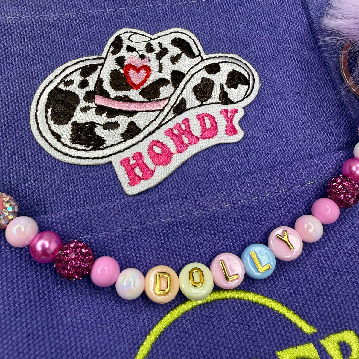 A close-up of a purple fabric item featuring a white embroidered patch with a cowgirl hat design, pink lettering reading 'HONDY', and a beaded necklace with pastel and glitter beads spelling 'DOLLY'. Dolly Parton Sweet but Strong mini tote by PrettyMUCH.