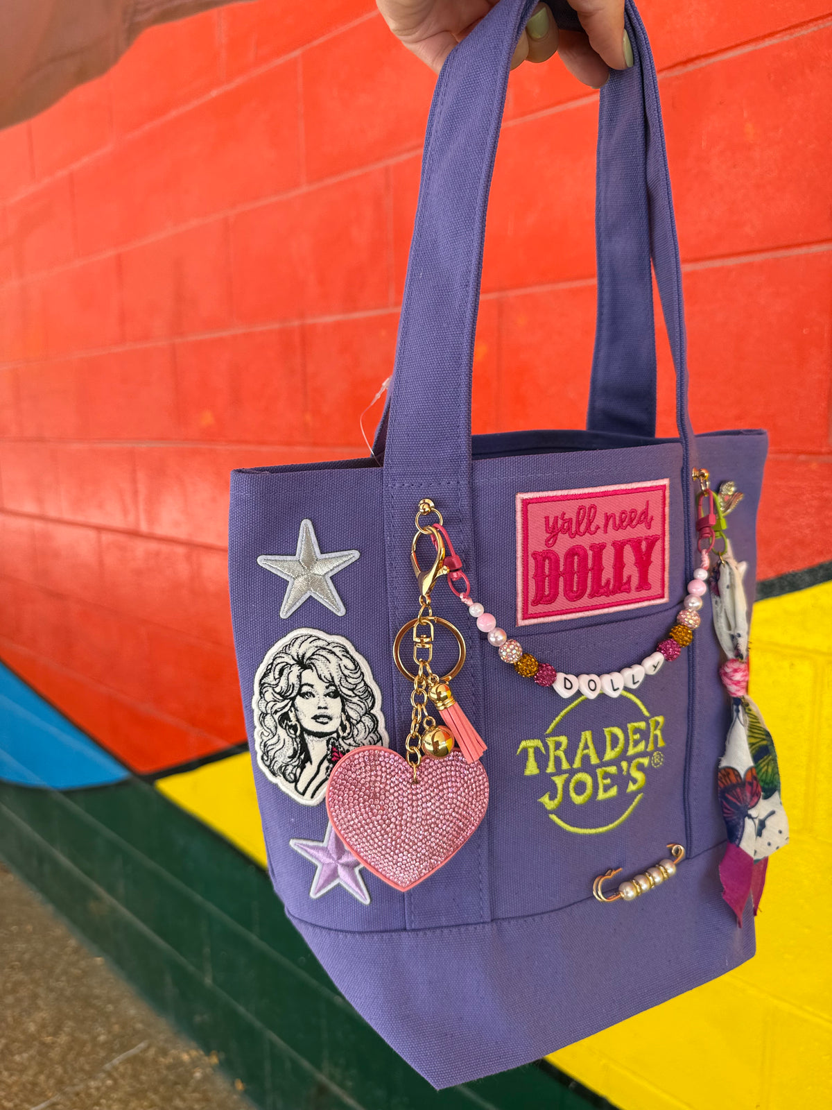 A purple Trader Joe's mini canvas tote decorated with Dolly patches, charms and keychains, held by a hand in front of a colorful red, yellow and green brick wall, with a hot pink 'y'all need DOLLY' patch, the green embroidered Trader Joe's logo and a pink rhinestone heart keychain. Dolly Parton Y'all Need Dolly mini tote by PrettyMUCH.