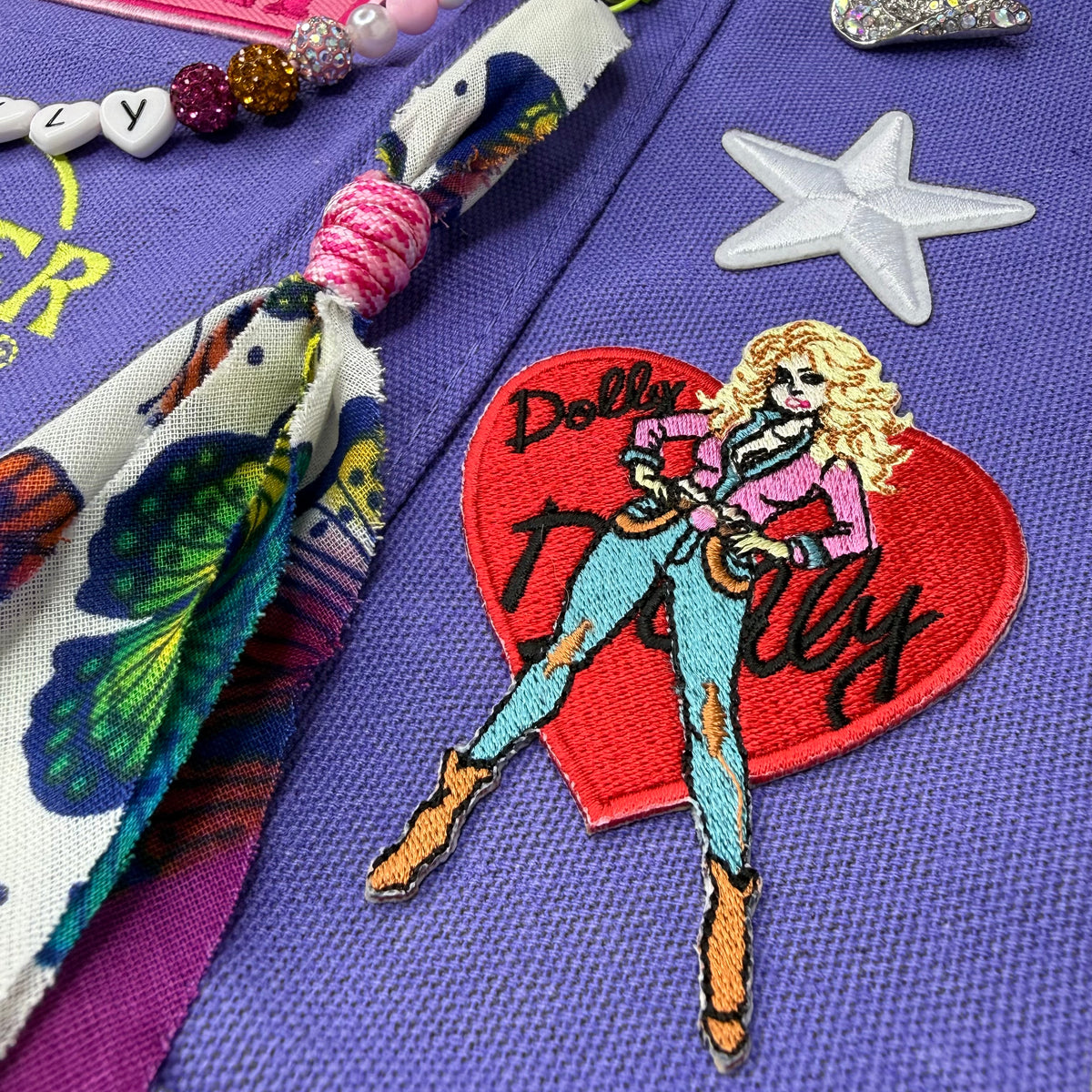 A close-up of a purple woven fabric surface decorated with colorful embroidered patches and accessories, including a red heart patch with a cartoon woman and the word 'Dolly' and a white star patch. Dolly Parton Y'all Need Dolly mini tote by PrettyMUCH.