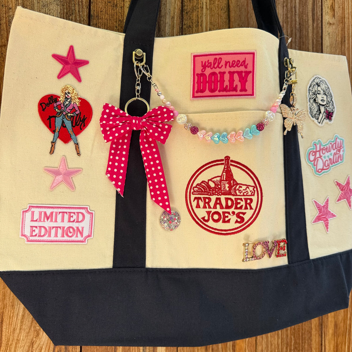 Dolly Parton Big Love large tote by PrettyMUCH, a natural and navy Trader Joe's canvas tote with all three Dolly patches, a beaded bag chain and a hot pink polka-dot bow.