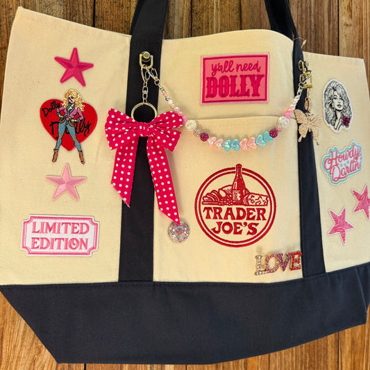 Dolly Parton Big Love large tote by PrettyMUCH, a natural and navy Trader Joe's canvas tote with all three Dolly patches, a beaded bag chain and a hot pink polka-dot bow.