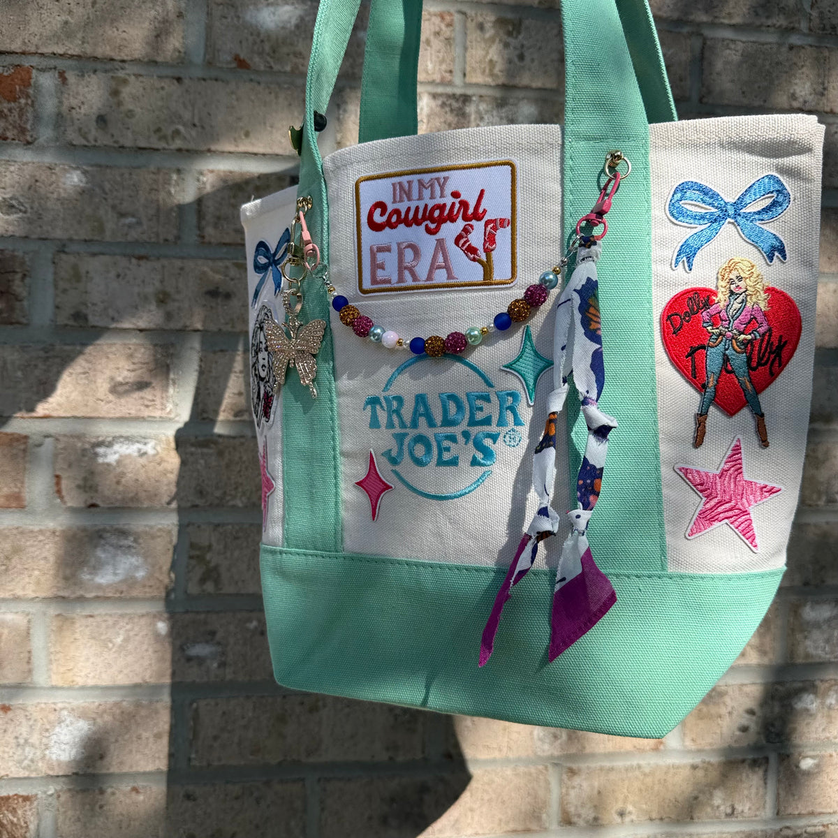 A mint green and cream canvas tote bag with various colorful embroidered patches, a beaded charm strap, and a 'Trader Joe's' patch, hanging against a brick wall. Dolly Parton Cowgirl Era mini tote by PrettyMUCH.