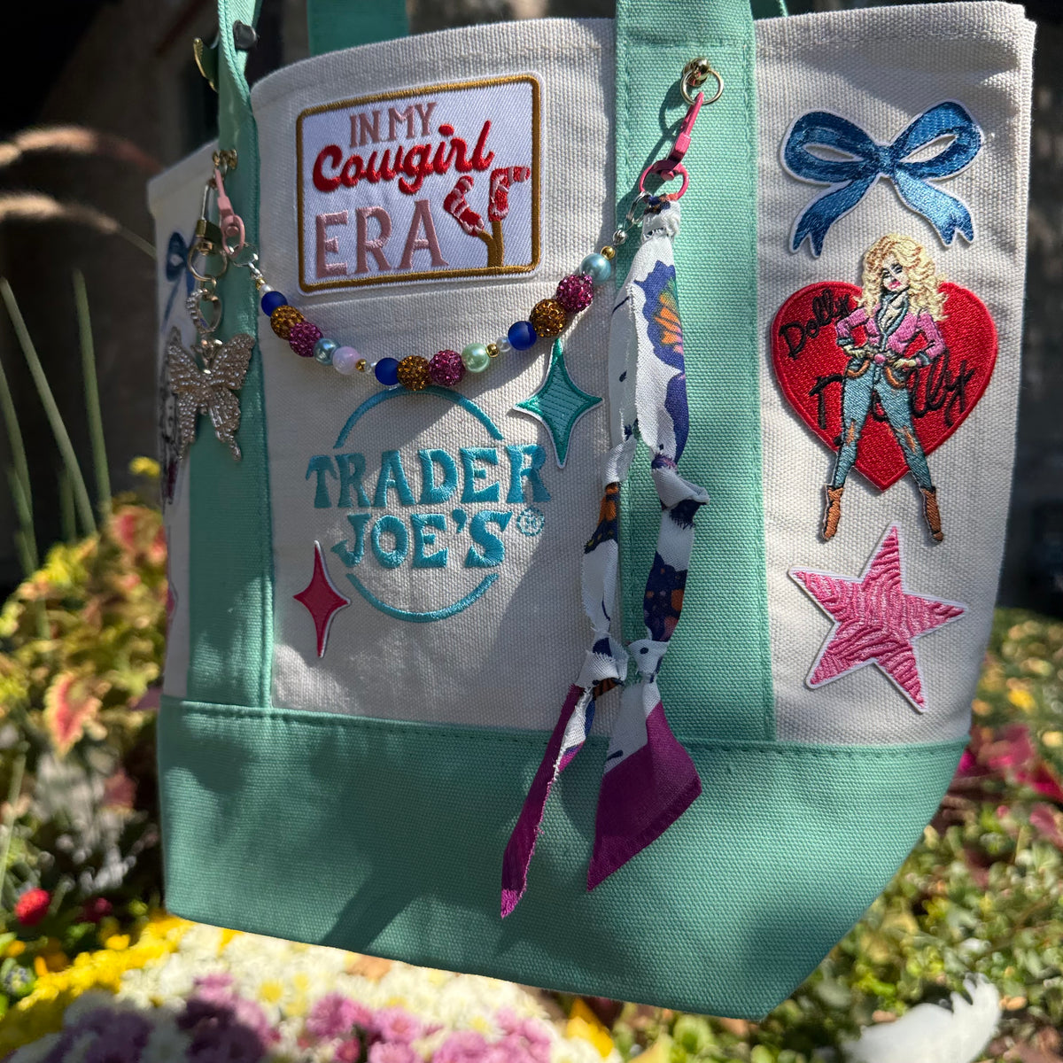 A canvas tote bag with mint green trim and multiple colorful embroidered patches and bead accessories, photographed outdoors in front of blurred flowers and foliage. Dolly Parton Cowgirl Era mini tote by PrettyMUCH.