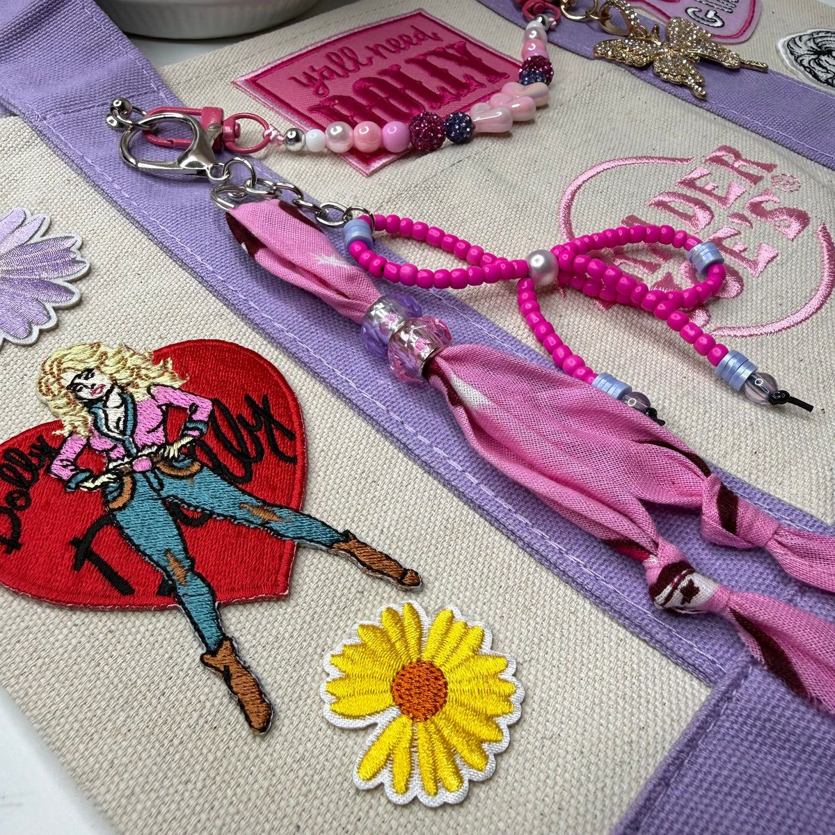 Close-up of the lavender and cream Trader Joe's mini tote with a pink beaded bow charm, a yellow daisy patch, the Dolly Parton red heart patch and a You Go Girl patch. Dolly Parton You Go Girl mini tote by PrettyMUCH.