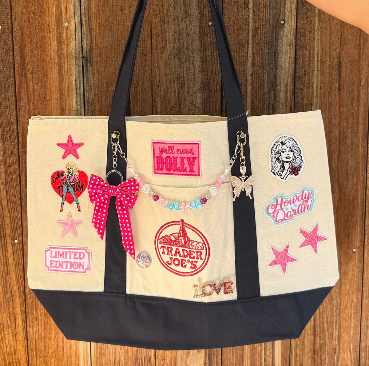 Dolly Parton Big Love large tote by PrettyMUCH, a natural and navy Trader Joe's canvas tote with all three Dolly patches, a beaded bag chain and a hot pink polka-dot bow.