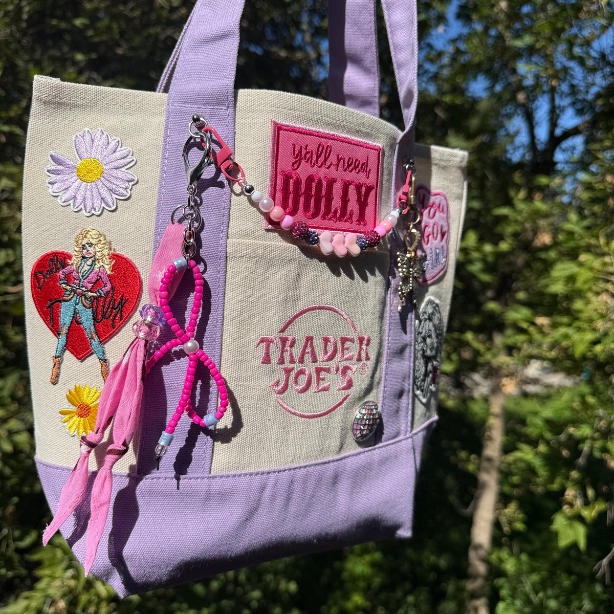 A lavender and cream Trader Joe's mini tote with embroidered patches, a pink beaded bag chain and a pink bandana twilly, photographed outdoors against green trees and blue sky. Dolly Parton You Go Girl mini tote by PrettyMUCH.