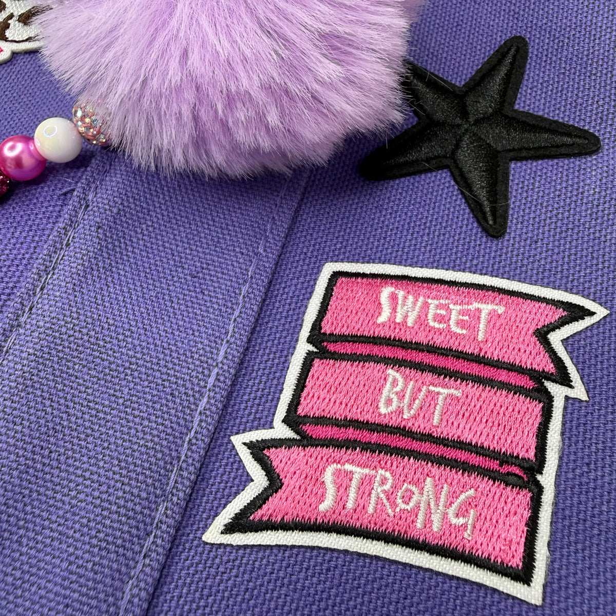 Close-up of a purple fabric item featuring a pink embroidered patch with the words 'SWEET BUT STRONG' in white, a black star applique, and a light purple fluffy pom-pom charm. Dolly Parton Sweet but Strong mini tote by PrettyMUCH.