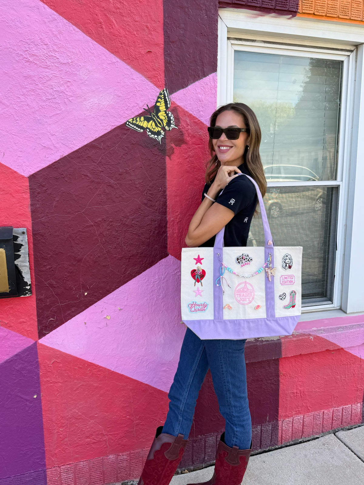 A woman wearing sunglasses, a black t-shirt, blue jeans and red boots stands in front of a colorful geometric wall, carrying a lavender and cream Trader Joe's large tote decorated with Dolly and western patches. Dolly Parton Howdy Darlin' large tote by PrettyMUCH.