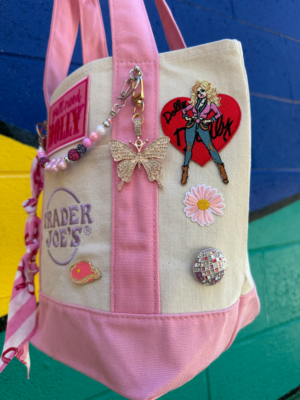 A pink and cream canvas tote bag decorated with various patches, pins, and charms, including a Trader Joe's logo patch, a butterfly charm, and a heart-shaped pin, set against a colorful blue and green background. Dolly Parton Pink Bandana mini tote by PrettyMUCH.