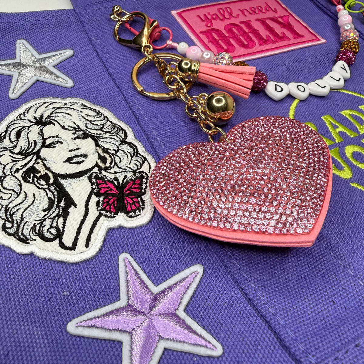 A pink rhinestone-embellished heart-shaped keychain bag with a pink tassel and gold-tone keyring, displayed on a purple embroidered fabric surface with decorative patches. Dolly Parton Y'all Need Dolly mini tote by PrettyMUCH.
