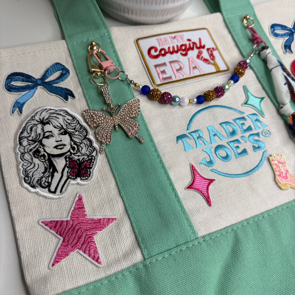 Close-up of the mint and cream Trader Joe's mini tote with a blue bow patch, the black-and-white Dolly Parton portrait patch with a pink butterfly, a pink zebra-stripe star and the embroidered Trader Joe's logo. Dolly Parton Cowgirl Era mini tote by PrettyMUCH.