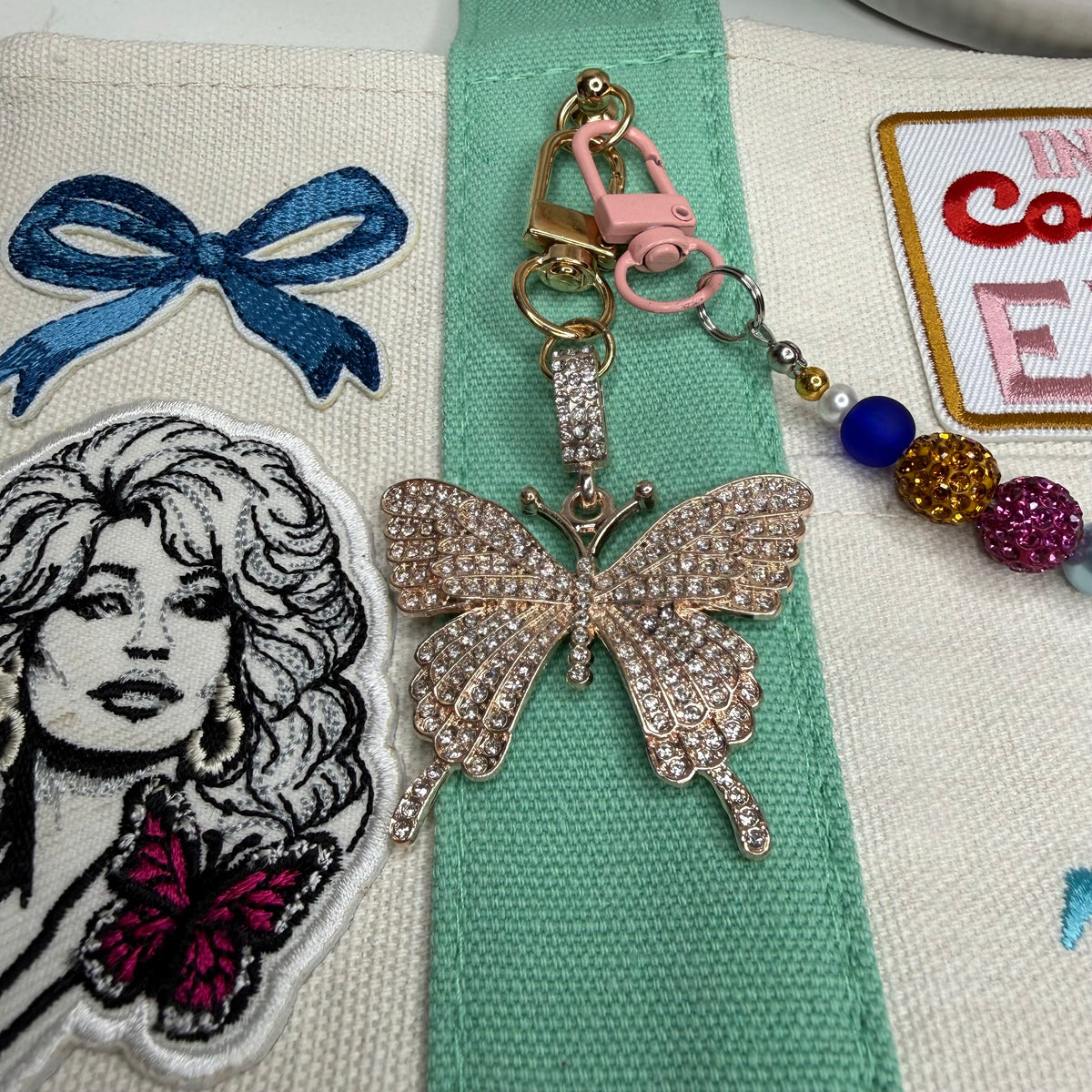 A rhinestone-embellished butterfly keychain charm with a pink lobster clasp and decorative beads, displayed on a beige embroidered fabric surface with other patches nearby. Dolly Parton Cowgirl Era mini tote by PrettyMUCH.