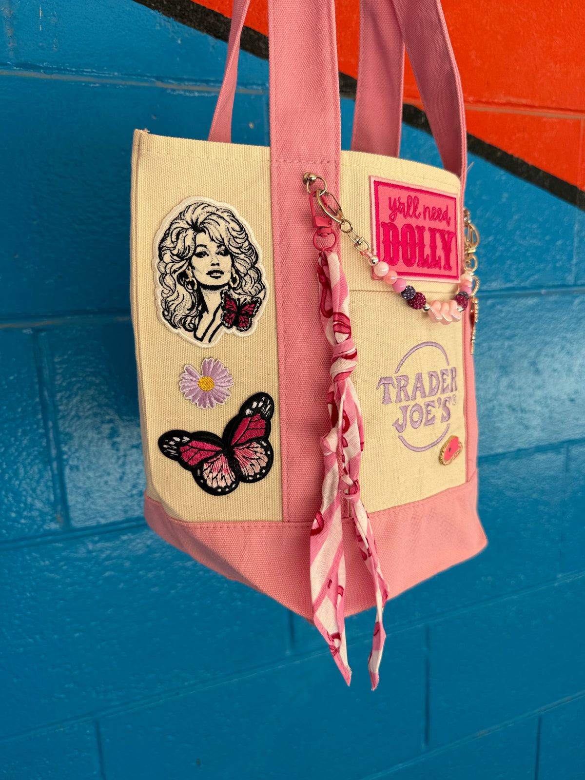 A pink and beige canvas tote bag with embroidered patches, a butterfly applique, and a pink fabric strap, hanging against a blue brick wall. Dolly Parton Pink Bandana mini tote by PrettyMUCH.