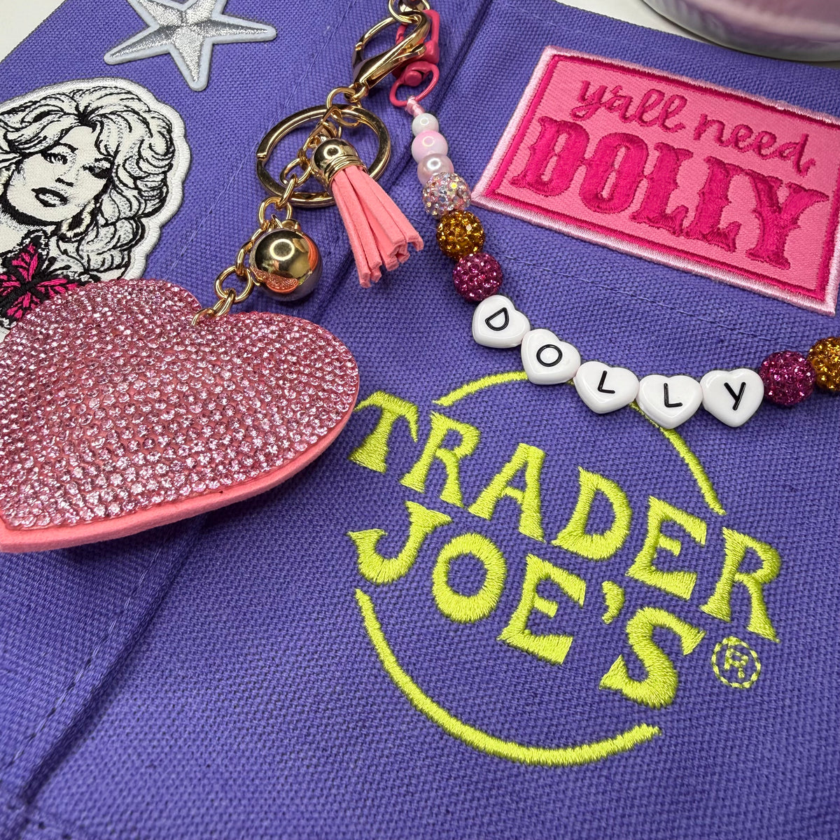 A close-up of a purple fabric surface with embroidered Trader Joe's branding, a pink rhinestone heart charm, a beaded keychain with a pink tassel, and decorative patches including one that reads 'y'all need BOLLY' and another with 'DOLLY' letter tiles. Dolly Parton Y'all Need Dolly mini tote by PrettyMUCH.
