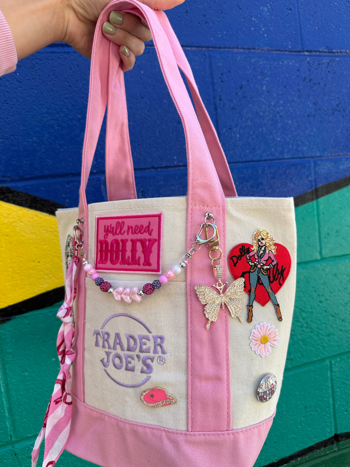 A hand holds a pink and cream canvas tote bag decorated with patches, pins, and charms, set against a colorful painted brick wall. Dolly Parton Pink Bandana mini tote by PrettyMUCH.