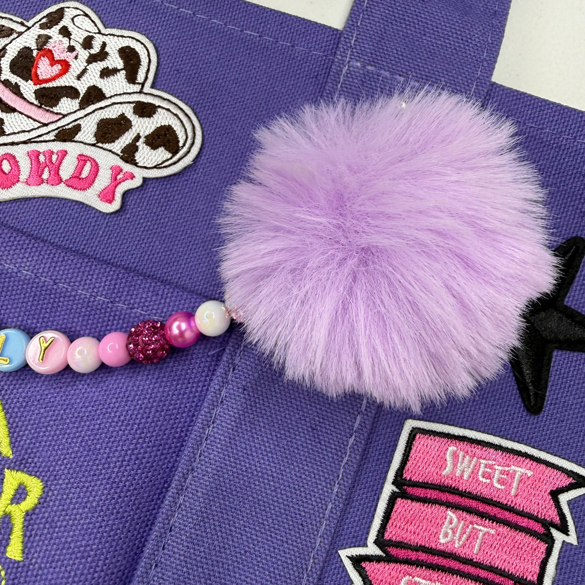 A close-up of a purple fabric tote bag decorated with embroidered patches, including a pink and white cow patch, a pink and white sweet but patch, and a beaded charm with a fluffy light purple pom-pom. Dolly Parton Sweet but Strong mini tote by PrettyMUCH.