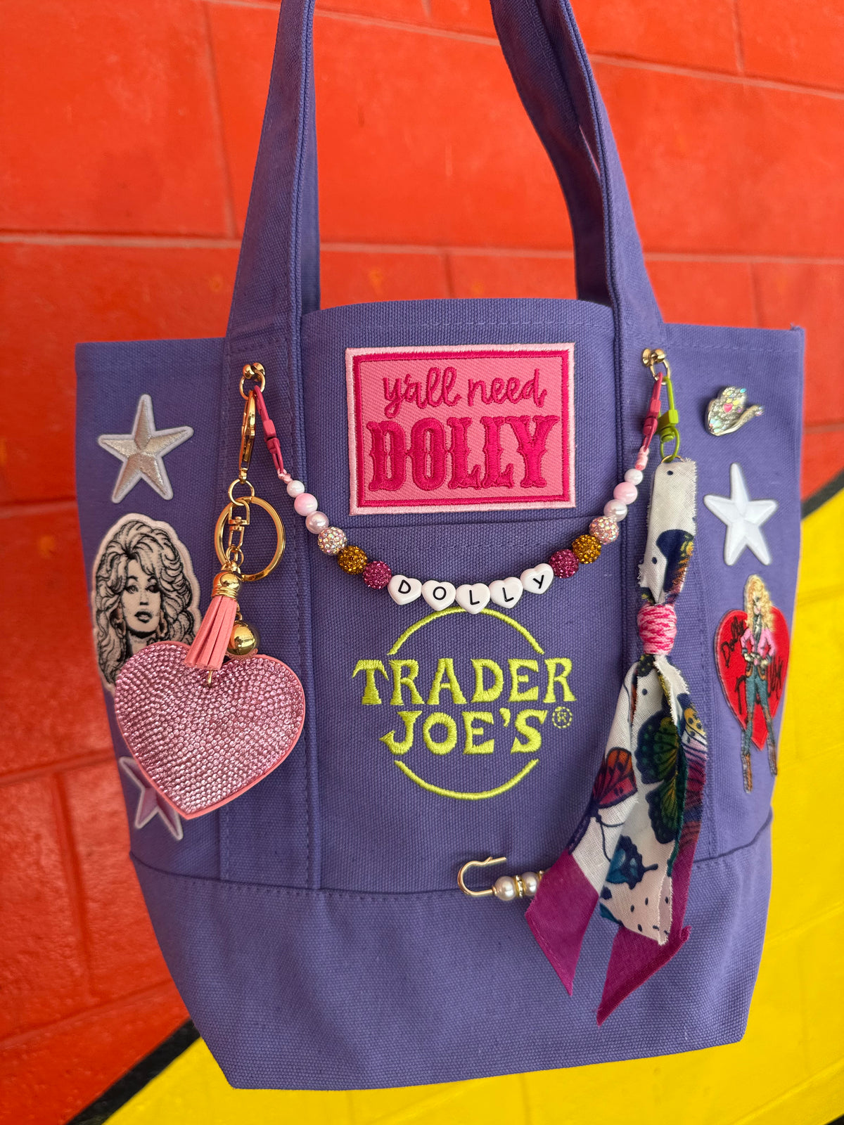 A purple canvas tote bag decorated with various pop culture patches, charms, and a beaded 'Dolly' necklace, set against a bright orange and yellow brick wall background. Dolly Parton Y'all Need Dolly mini tote by PrettyMUCH.