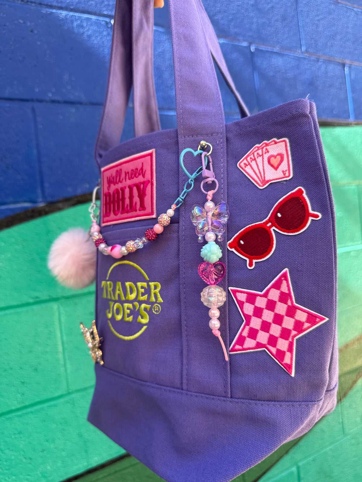 A purple canvas tote bag with various colorful patches, a beaded charm, and a pink pom-pom, shown against a blue and green painted brick wall. Dolly Parton Aces High mini tote by PrettyMUCH.