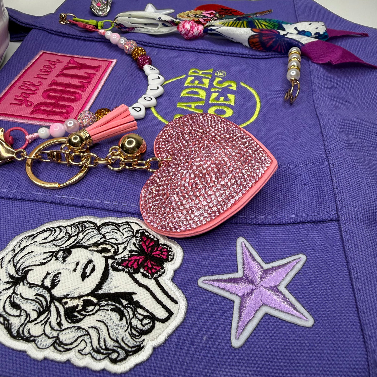 A close-up of a purple embroidered tote bag with decorative patches and accessories laid on top, including a pink rhinestone heart keychain, a tassel charm, letter beads spelling 'DOLLY', a star patch, and other colorful embroidered details. Dolly Parton Y'all Need Dolly mini tote by PrettyMUCH.