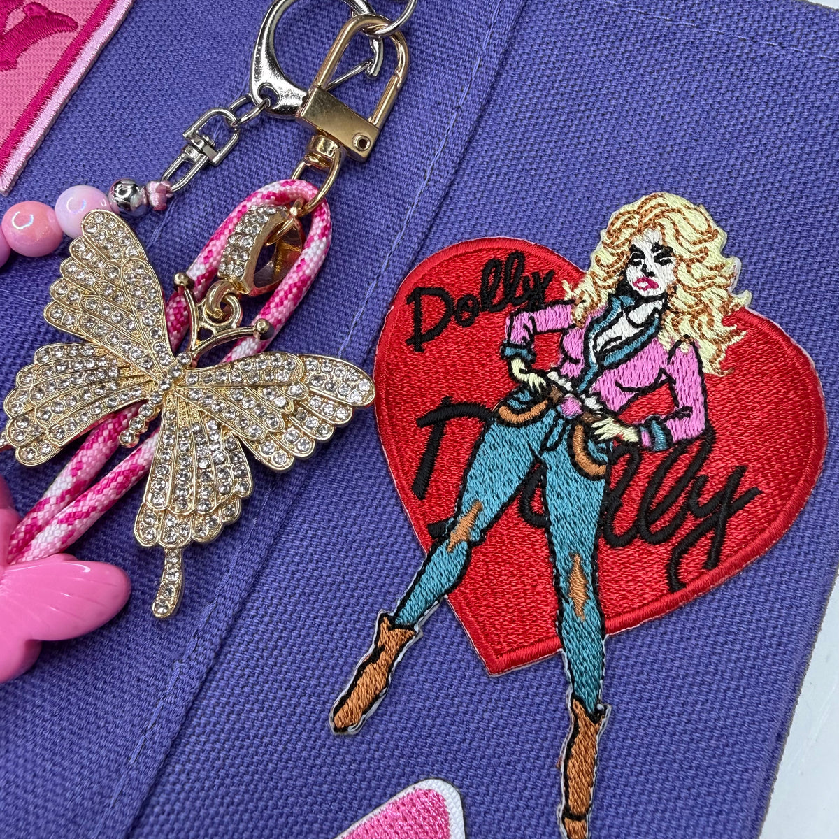 A close-up of a purple fabric accessory featuring a colorful embroidered Dolly Parton patch on a red heart, a gold rhinestone butterfly charm, pink beads, and a metal clasp. Dolly Parton Boots & Heels mini tote by PrettyMUCH.